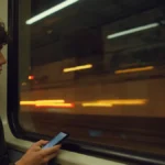 A young person with headphones looks out a subway window at night, with blurred city lights reflected in the glass.