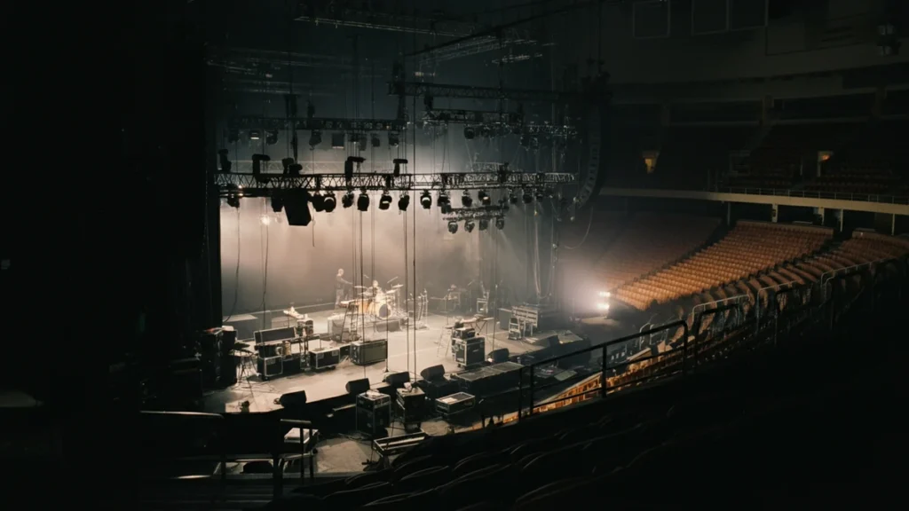 An empty arena stage being set up by a road crew under dramatic work lights, viewed from the seats.