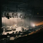 An empty arena stage being set up by a road crew under dramatic work lights, viewed from the seats.