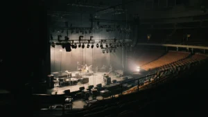 An empty arena stage being set up by a road crew under dramatic work lights, viewed from the seats.