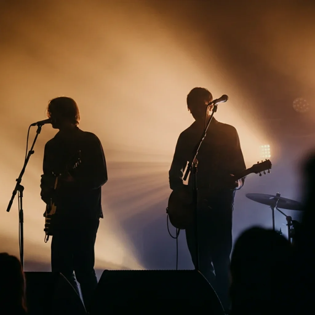 Silhouettes of a singer and guitarist performing on a smoky, backlit indie rock concert stage.