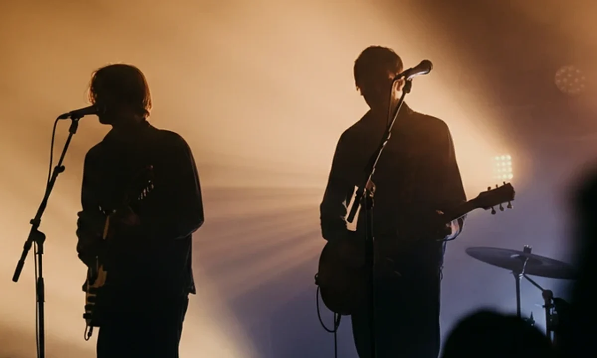 Silhouettes of a singer and guitarist performing on a smoky, backlit indie rock concert stage.