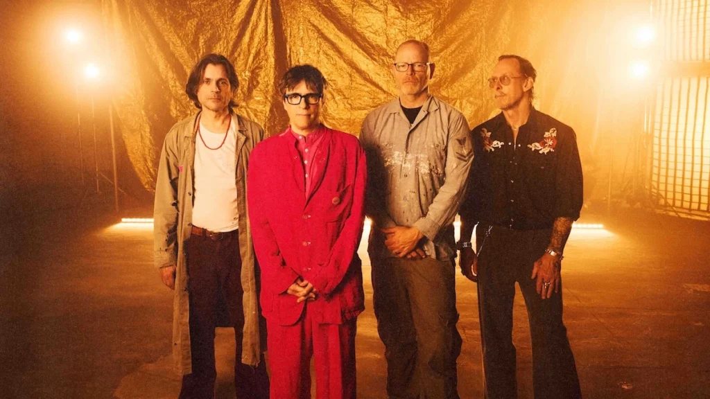 Weezer photographed against a gold backdrop for the Gold Album era, with Rivers Cuomo in a red suit at the centre of the four-piece