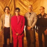 Weezer photographed against a gold backdrop for the Gold Album era, with Rivers Cuomo in a red suit at the centre of the four-piece